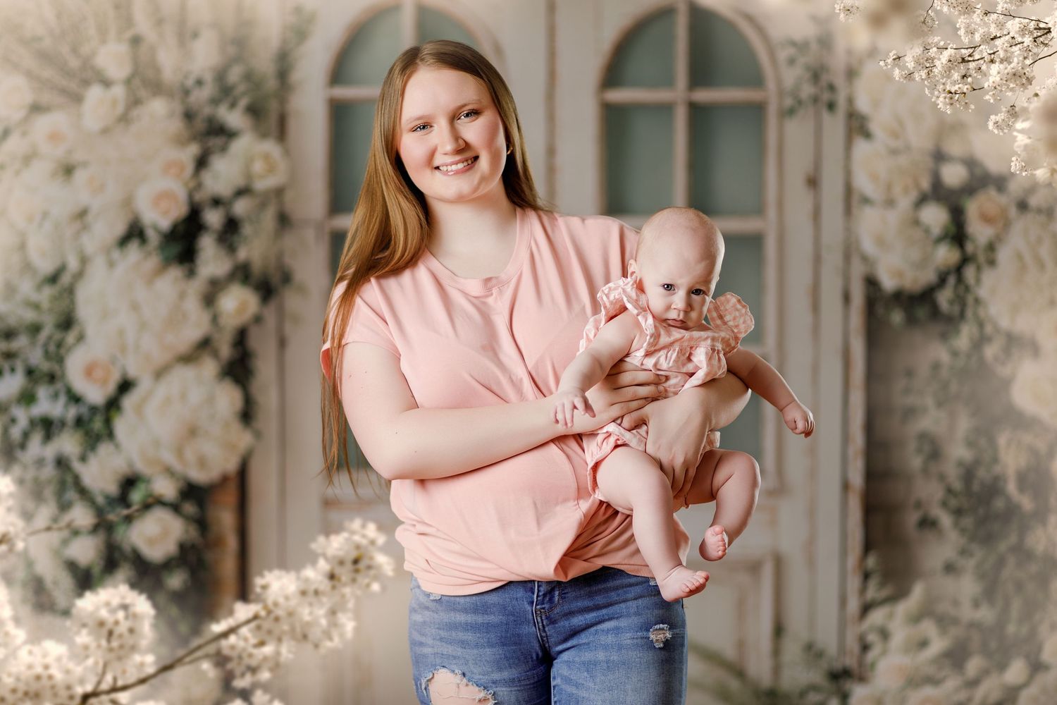 Young mother in pink top and jeans smiles holding baby in floral studio portrait.