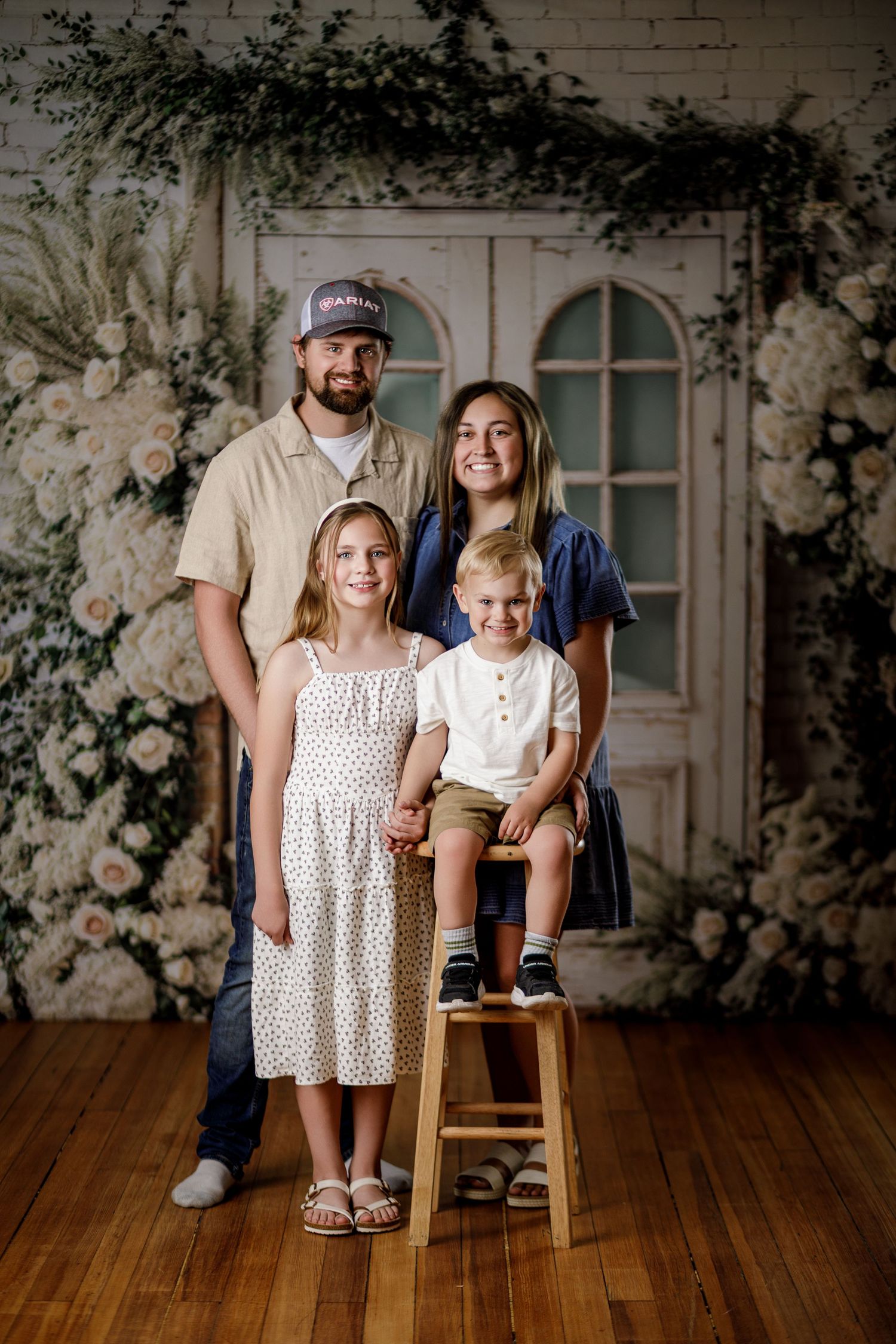 Family of four posing for portrait in front of floral backdrop with white roses and greenery on wooden floor.