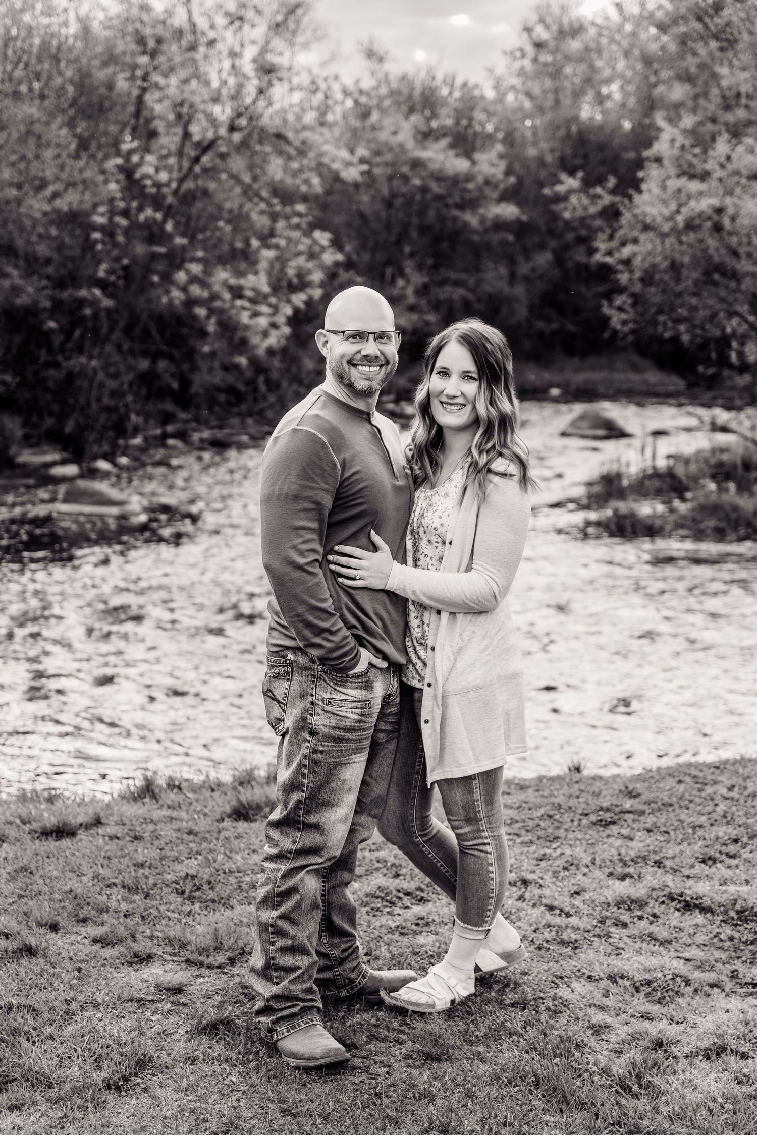 Black and white photo of a smiling couple embracing outdoors near a creek surrounded by lush trees.