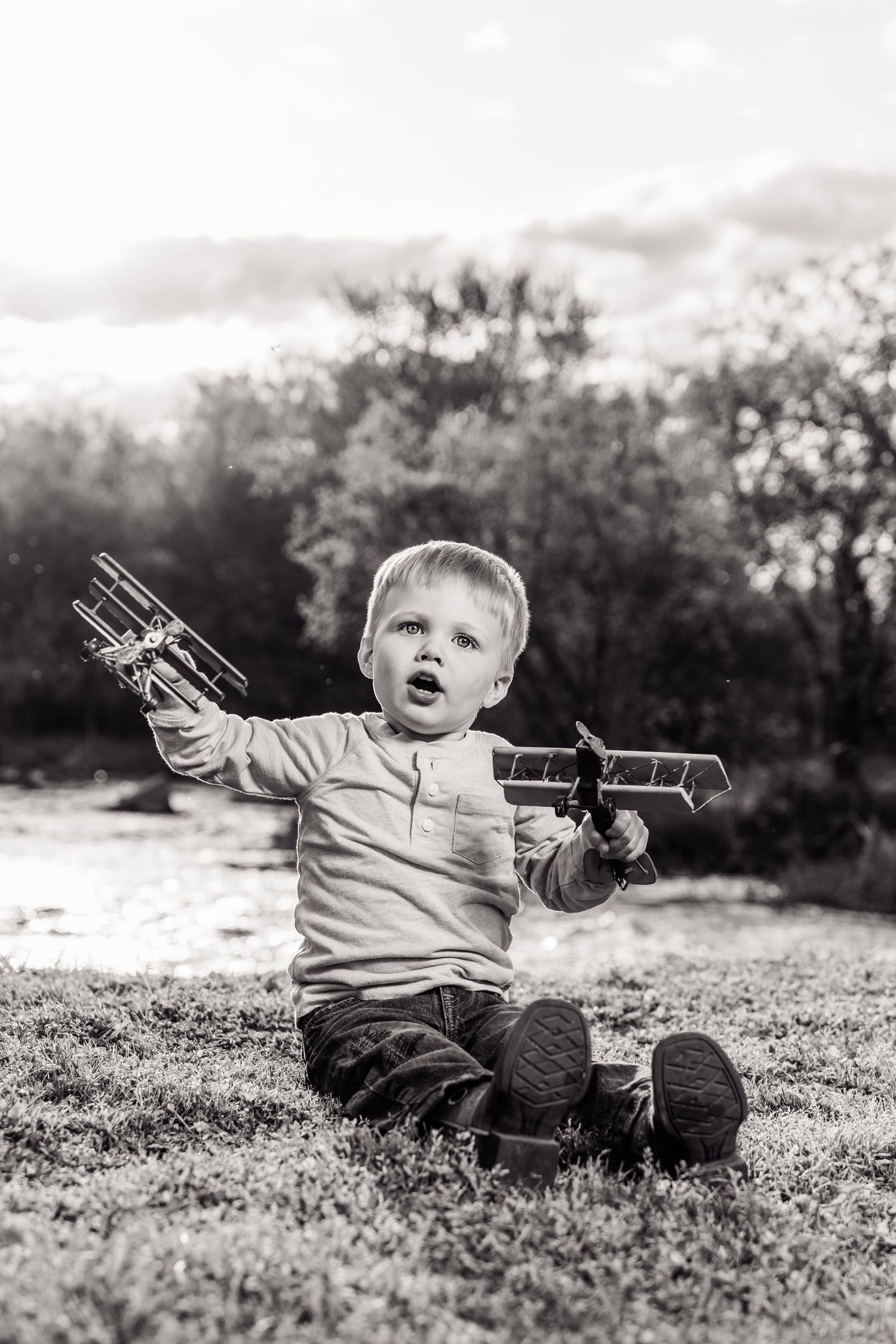 Black and white photo of a toddler boy sitting on grass holding two toy wooden airplanes with mouth open in wonder.