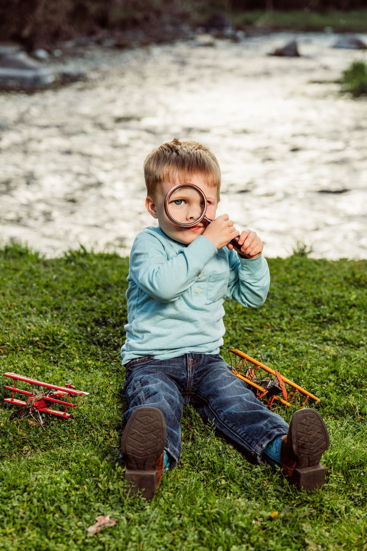 Young boy sitting on grass by a river, holding a magnifying glass up to his eye with toy planes nearby.