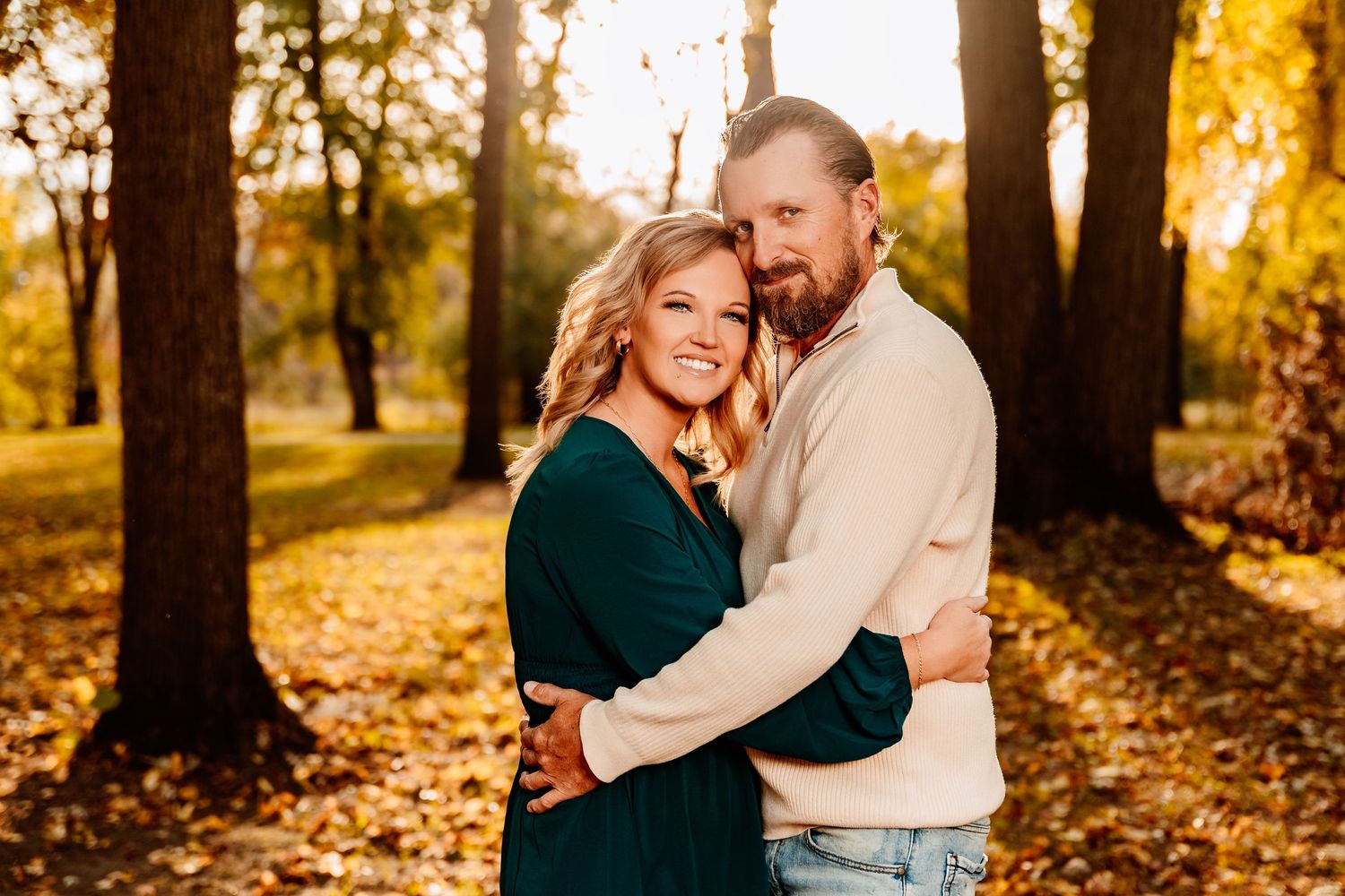 A couple embraces while laughing together in a sunlit autumn forest with golden leaves scattered on the ground.