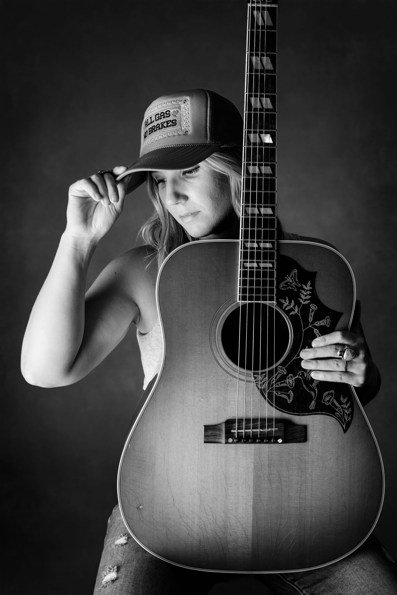 Black and white photo of woman tipping trucker hat while holding acoustic guitar in studio.