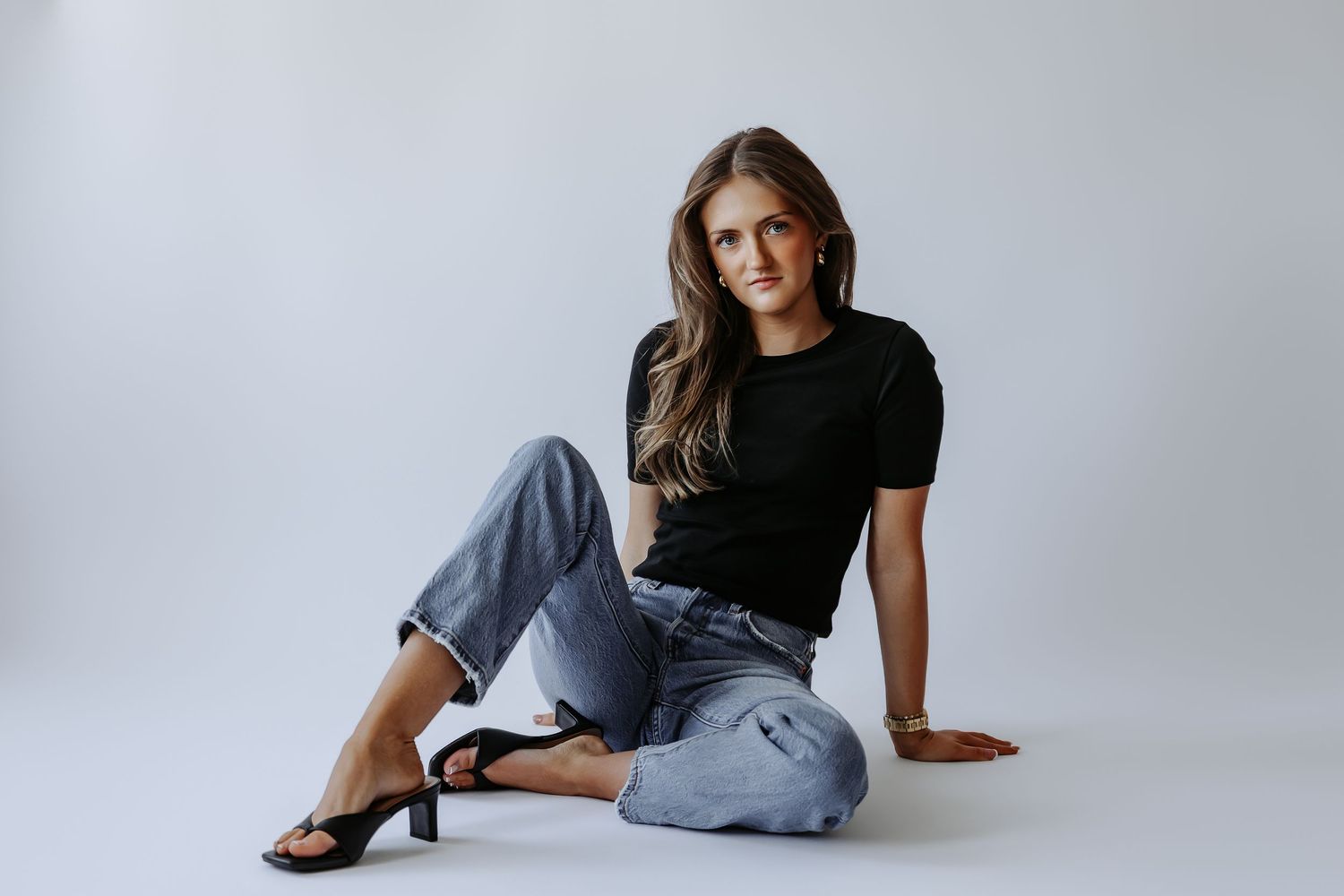 Woman in black tee and jeans sitting on white floor wearing black heeled sandals.