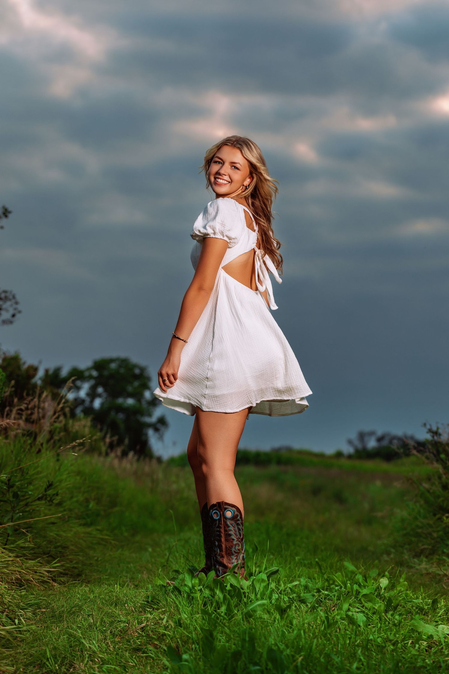 Smiling blonde woman in white dress and cowboy boots standing in green field under dramatic cloudy sky.