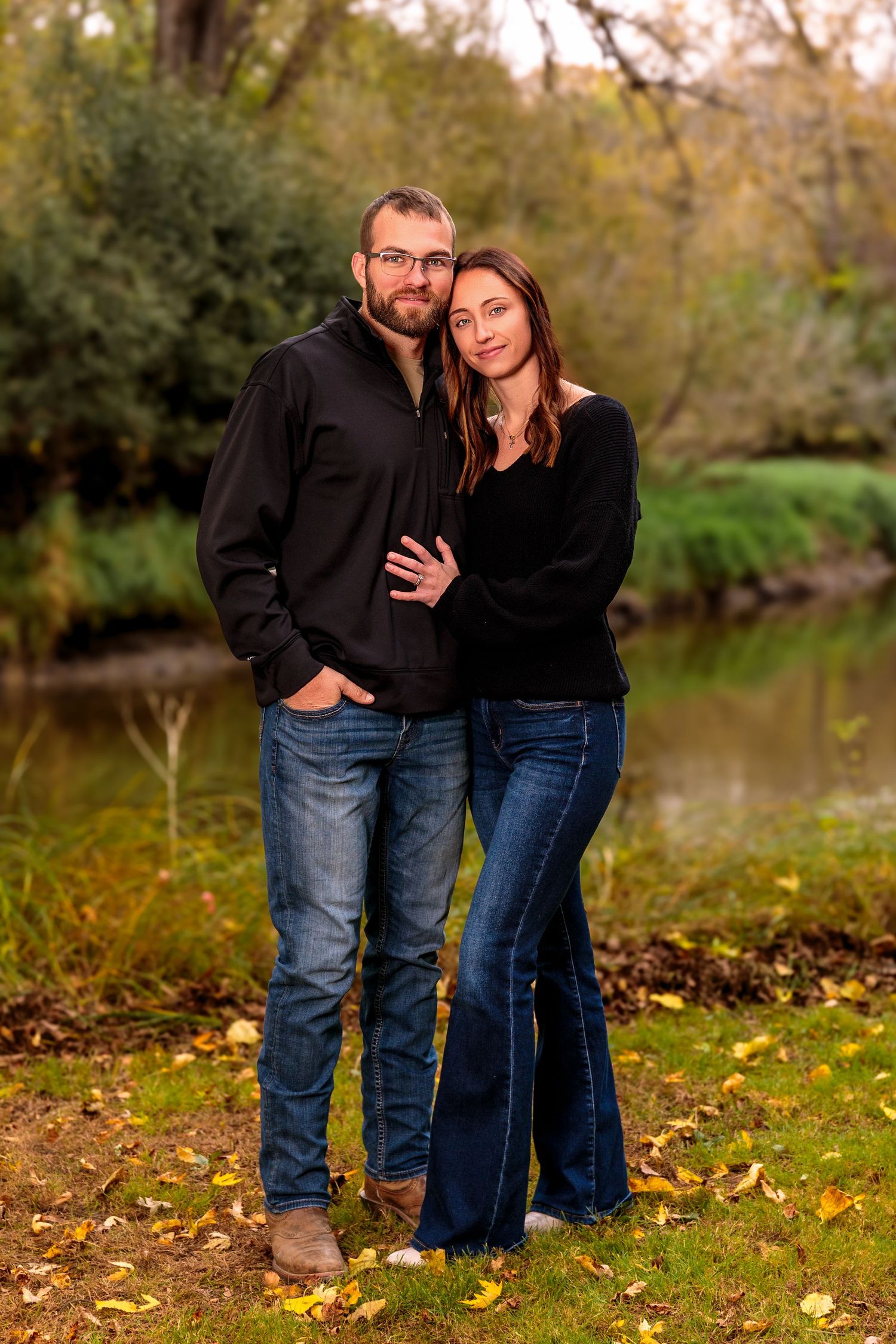 Romantic couple poses together on fallen leaves by waterfront during golden hour.