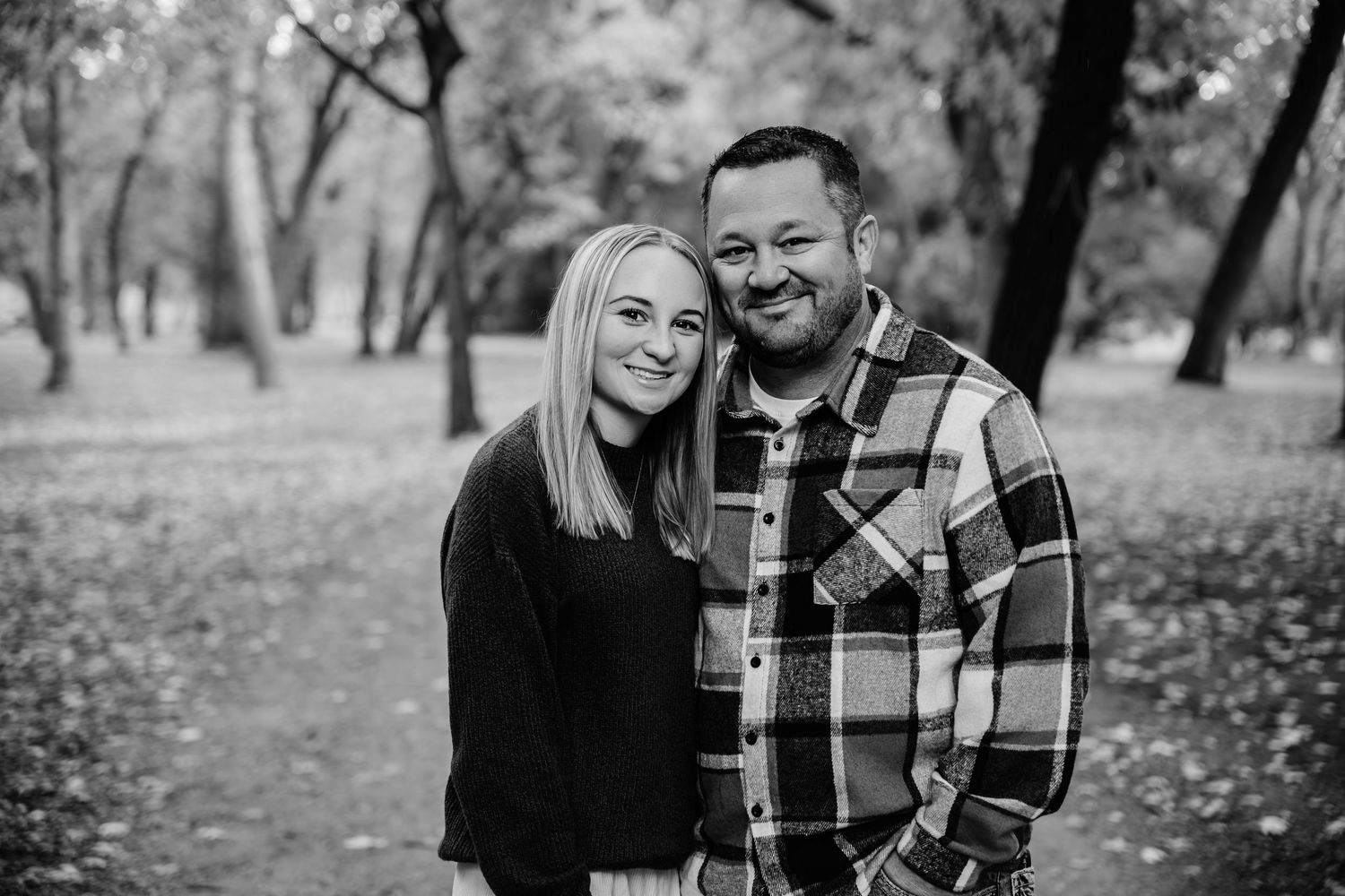 A couple poses together outdoors in a park during autumn, with trees and fallen leaves visible in the background.