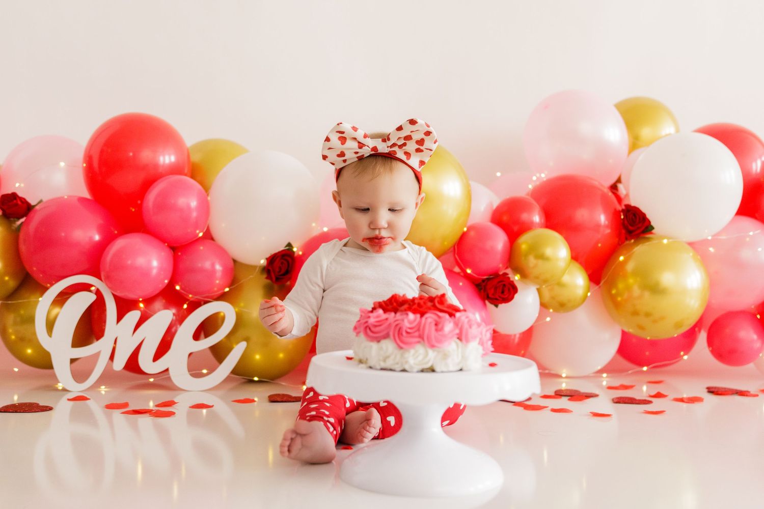 Pink and gold balloon garland with white 'One' sign for first birthday celebration.