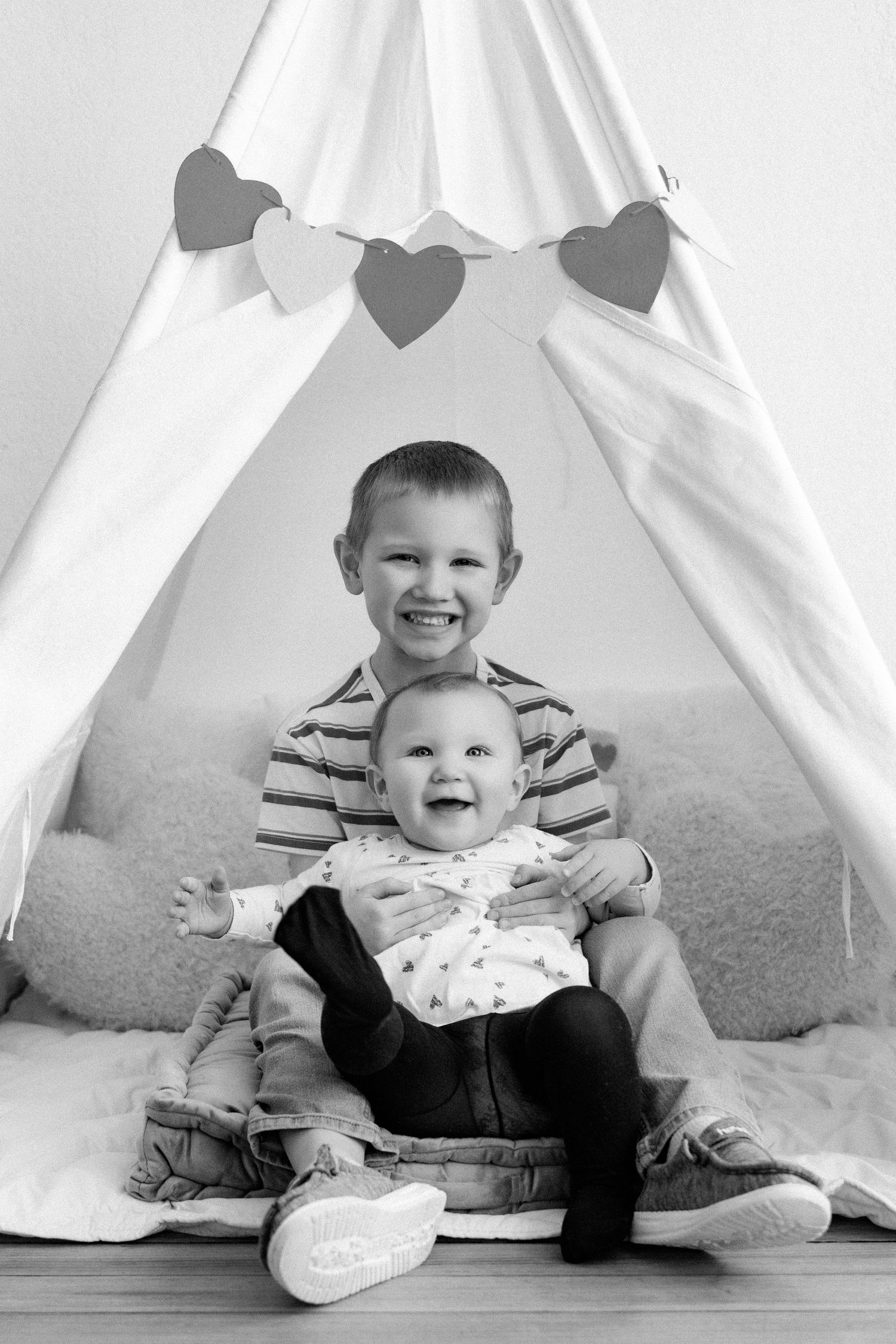 Two small children sitting together inside a white teepee tent on carpeted floor.