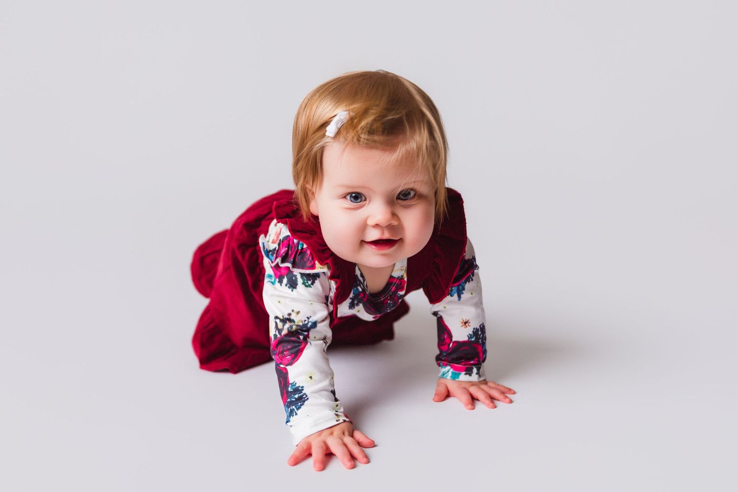 Smiling baby in burgundy outfit crawling on white studio floor.