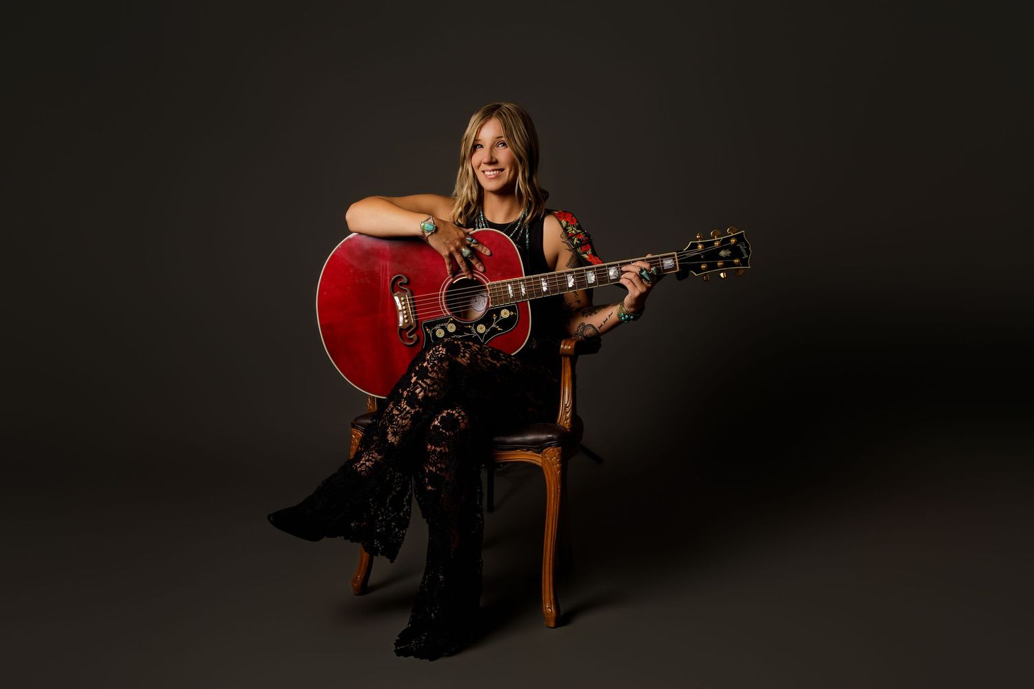 Smiling female guitarist sitting on chair playing red acoustic guitar against dark background in professional studio photo.