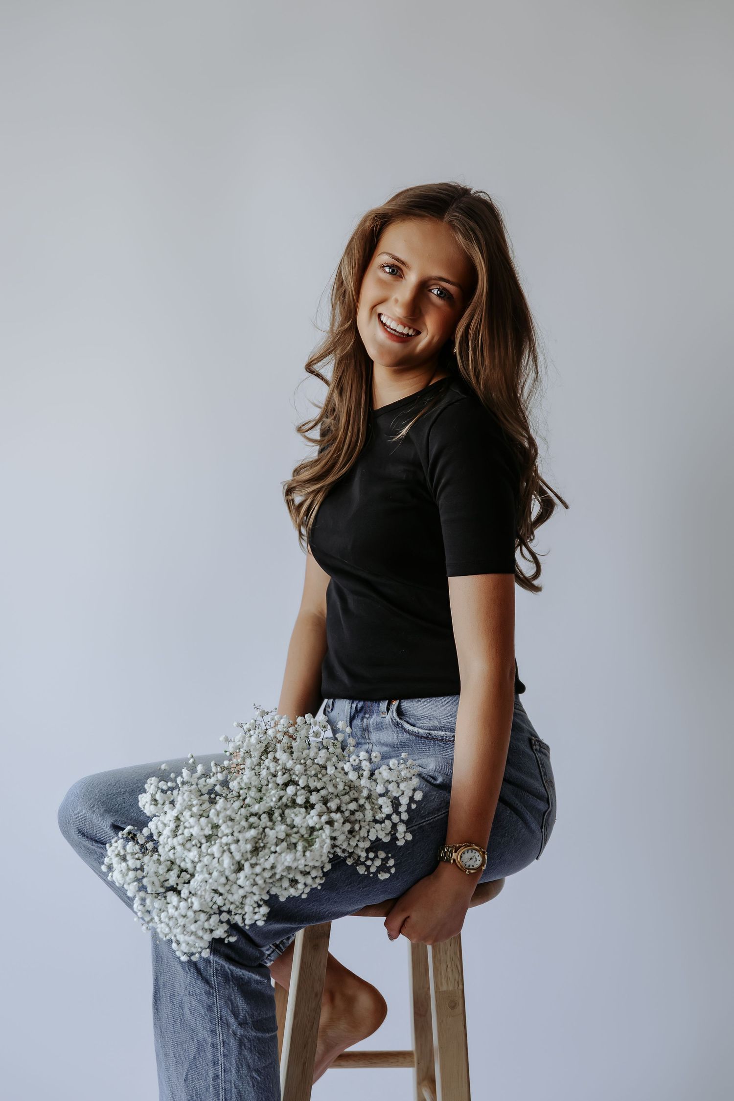 Smiling young woman in black tee and jeans holding white baby's breath flowers while seated on a wooden stool.