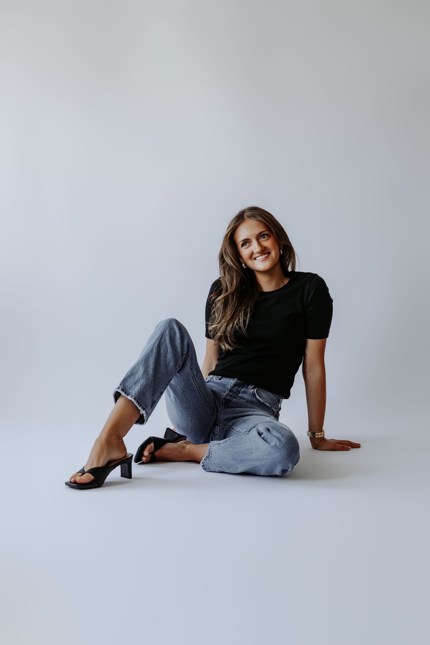 Smiling young woman sitting on floor in black tee and light blue jeans with black heeled sandals.