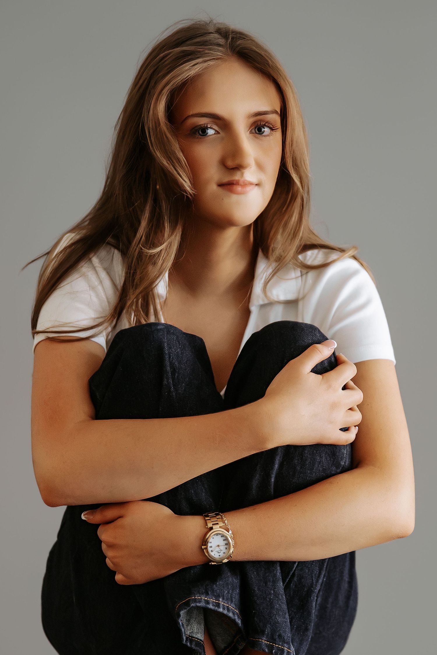 Young woman hugging knees wearing white t-shirt, gold watch, and dark jeans, studio portrait.