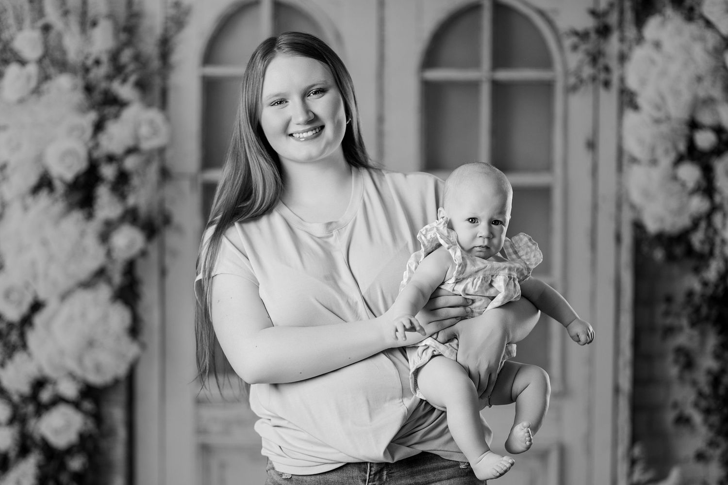 Black and white photo of a smiling young woman holding a baby in a floral studio setting.