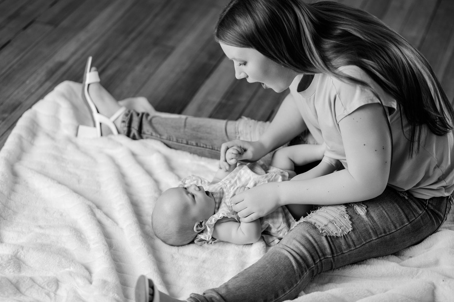 Black and white photo of a young mother sitting on a blanket interacting with her baby lying down.