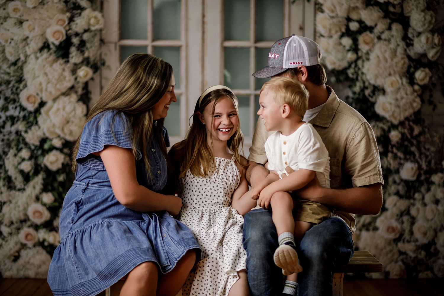 Family of four smiling together in front of a floral backdrop with white roses.
