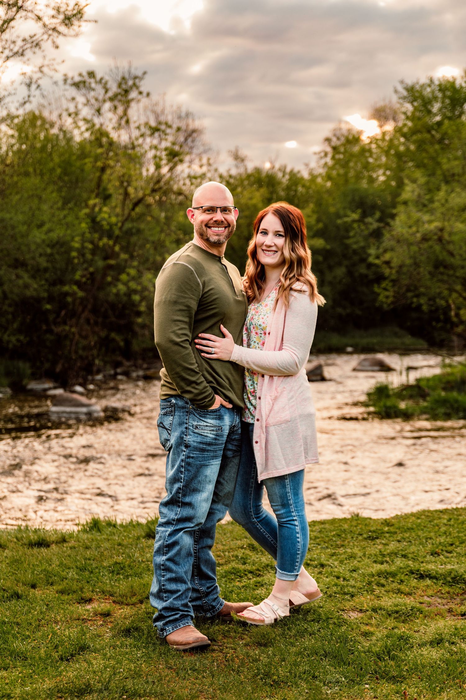 A smiling couple poses outdoors near a creek, man in green shirt and jeans, woman in pink cardigan and blue jeans.