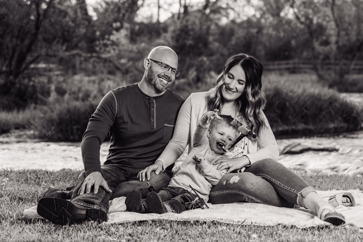 Black and white photo of a happy family with a laughing toddler sitting on a blanket outdoors.