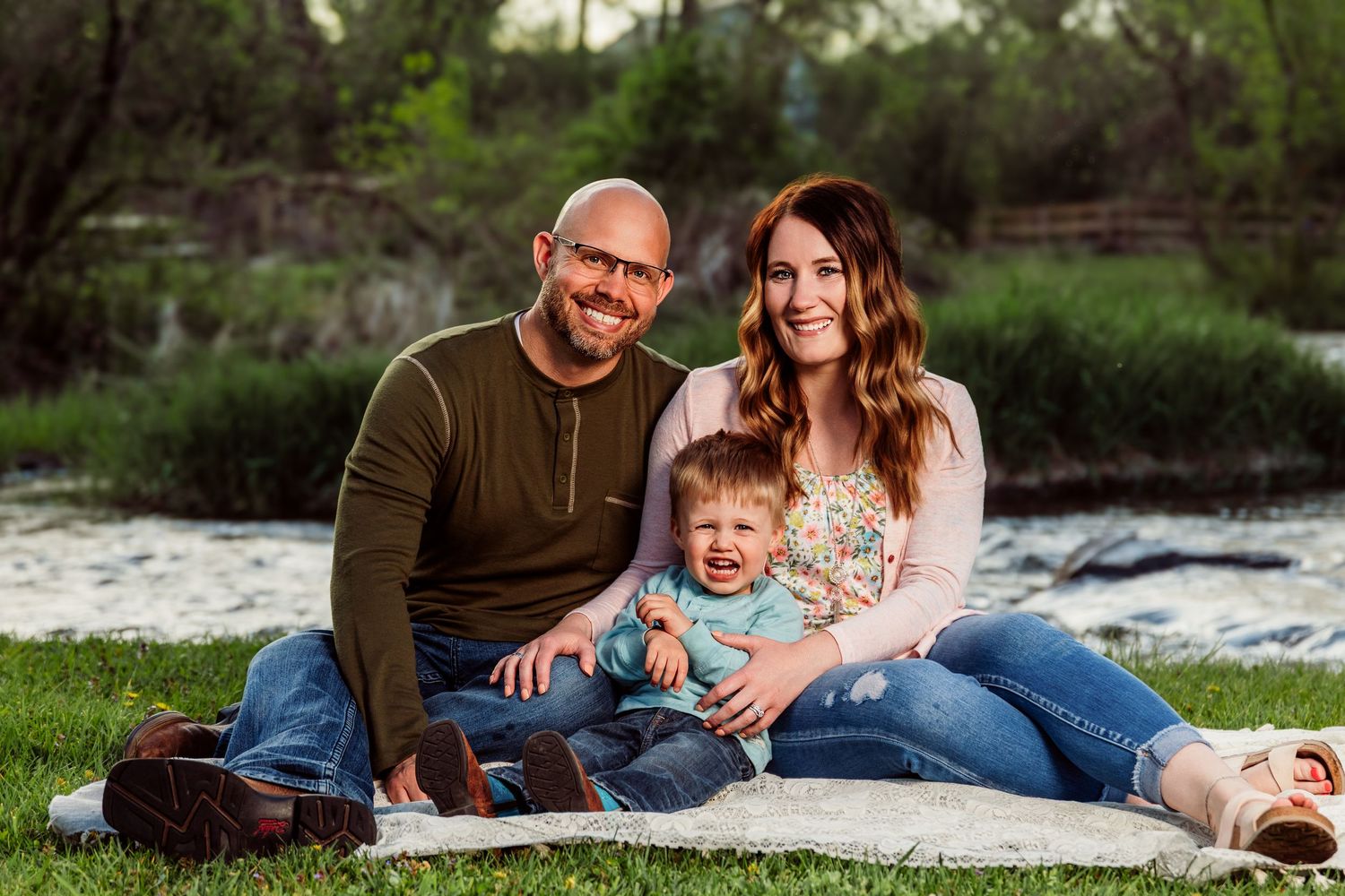 Happy family of three sitting on a blanket outdoors near a river, smiling parents with their baby boy.
