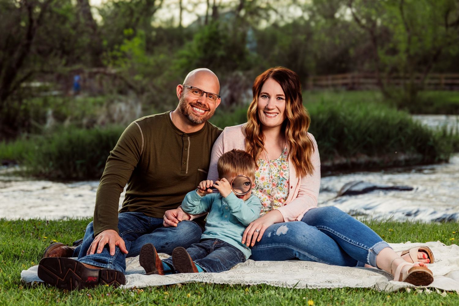 Happy family of three smiling outdoors on a blanket by a river in a lush green park setting.