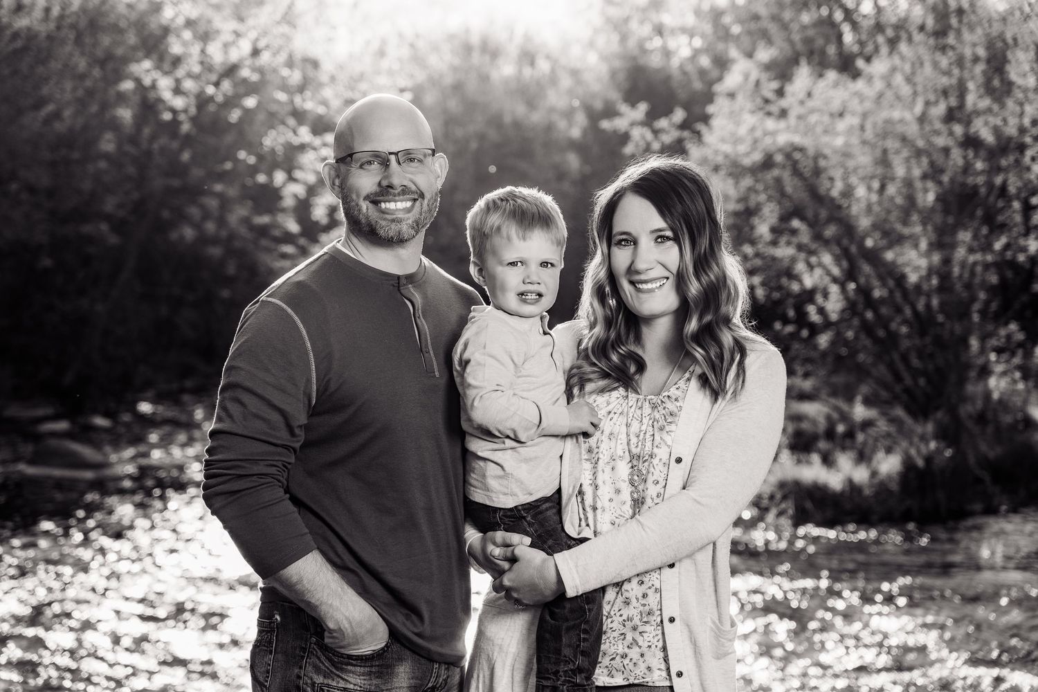 Black and white family portrait of a man, woman, and toddler boy smiling outdoors near trees and water.