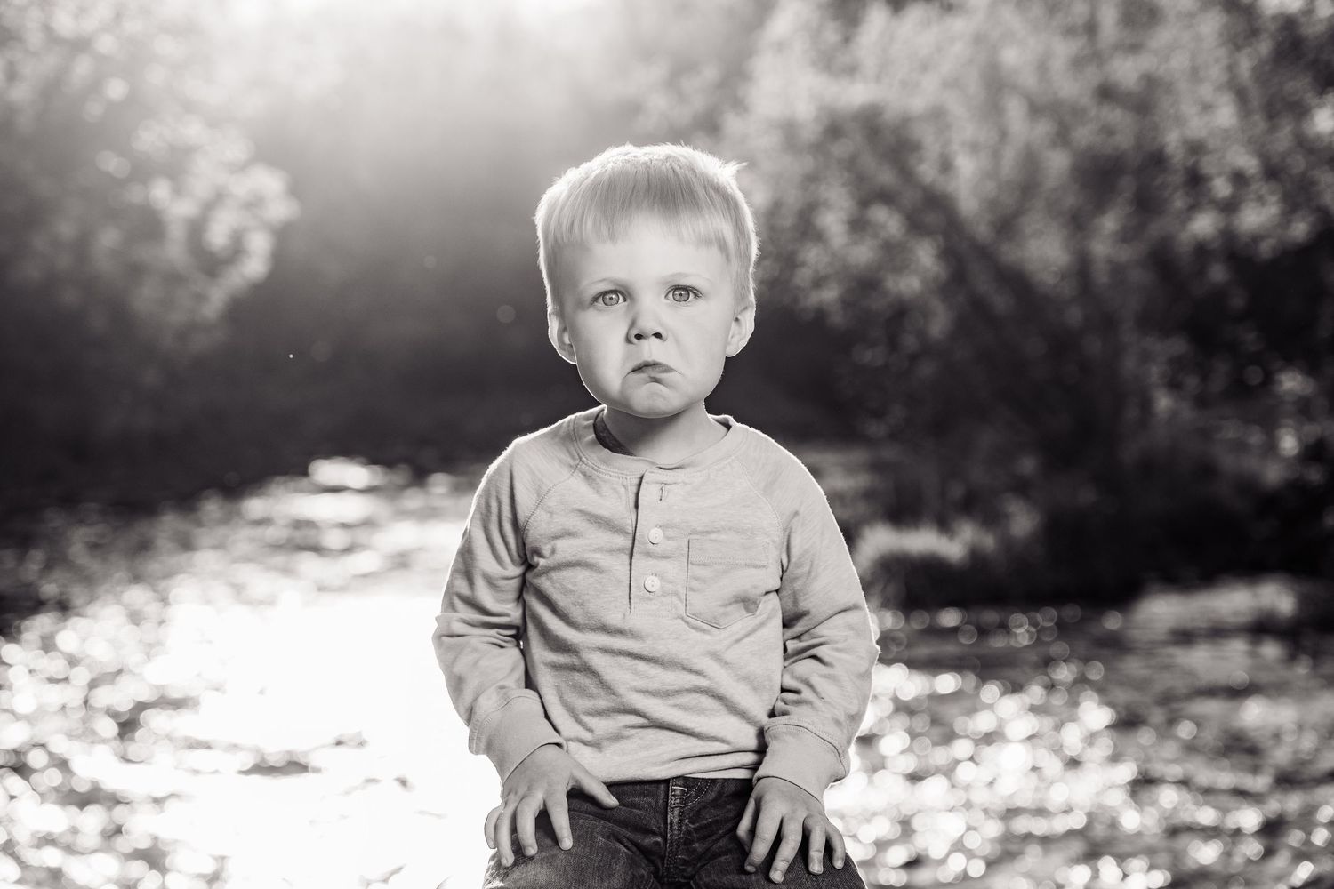 Black and white portrait of a young blond boy standing near a sunlit river, looking thoughtfully at the camera.