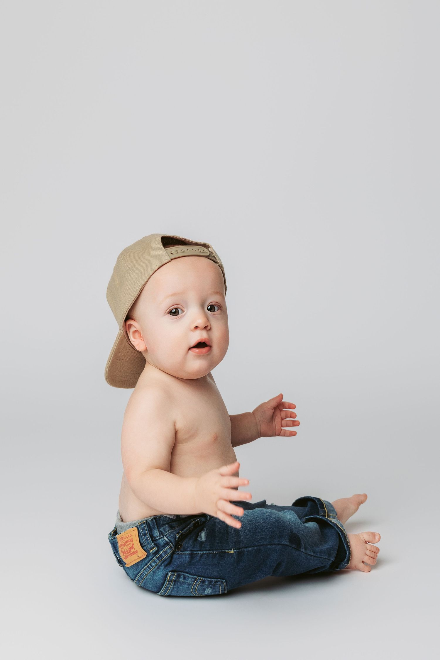 Cute shirtless baby boy sitting in blue jeans and a beige snapback cap on a light gray background.