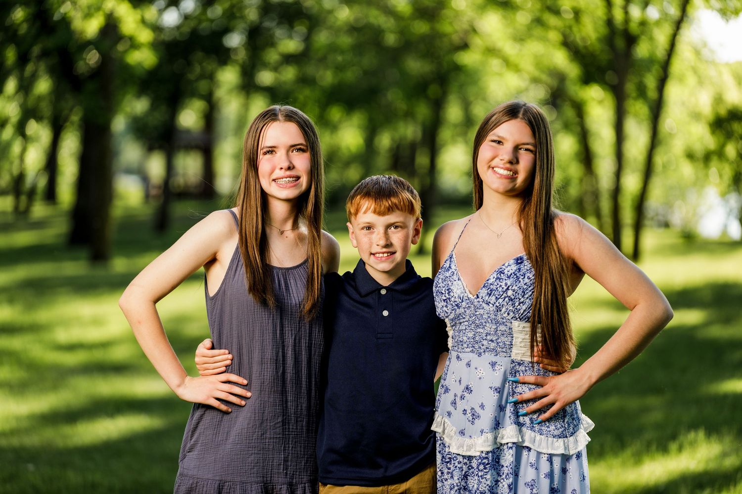 Two teenage girls and a young red-haired boy posing together in a sunny green park setting.