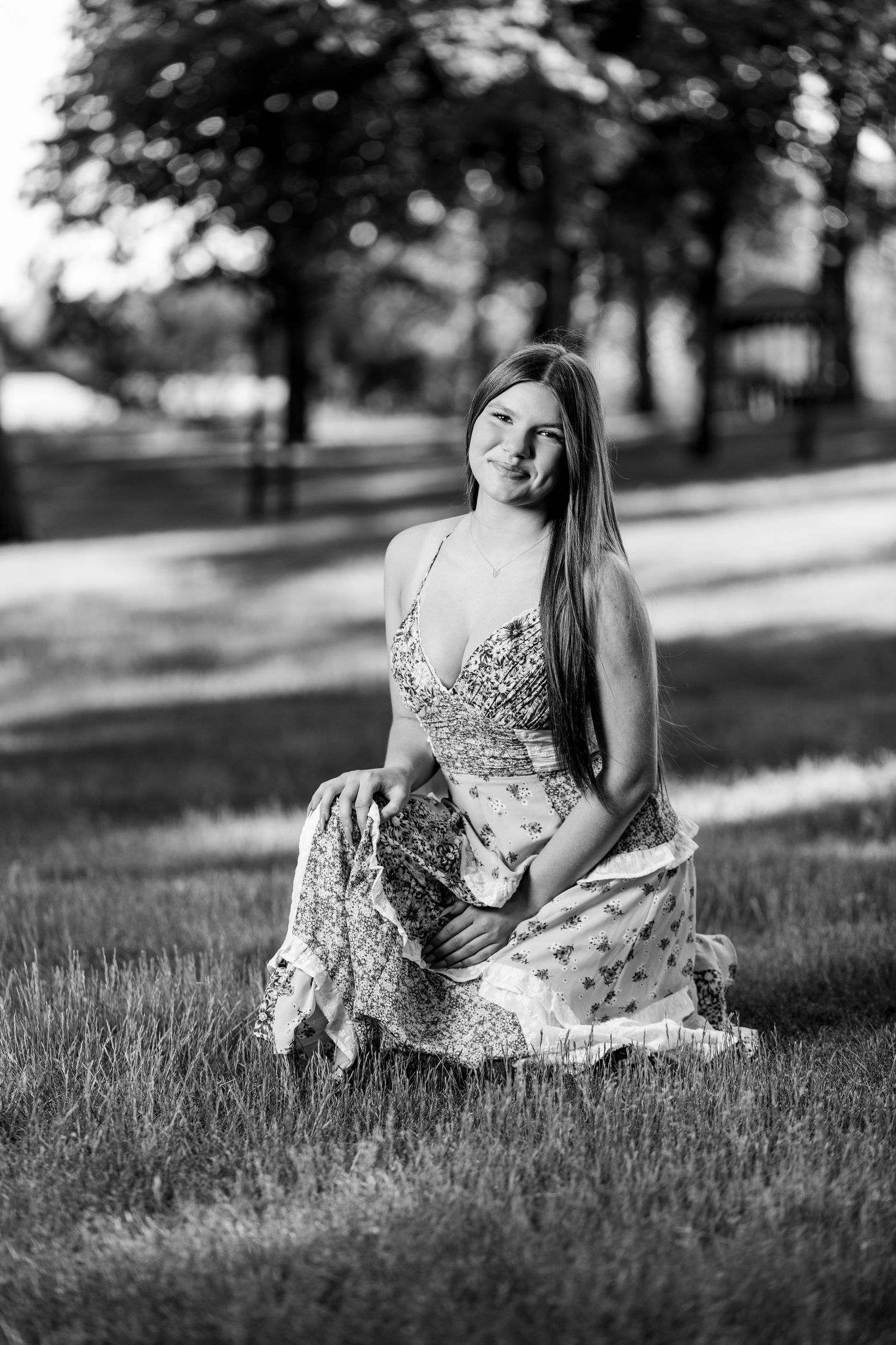 Young woman in a floral dress sitting gracefully on grass in a park, black and white photo.