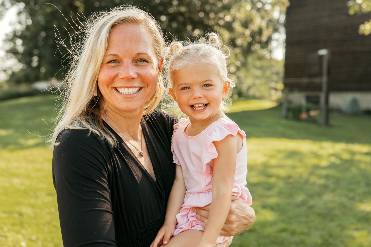 Woman in black shirt holding young child in pink outfit while standing in sunny backyard.