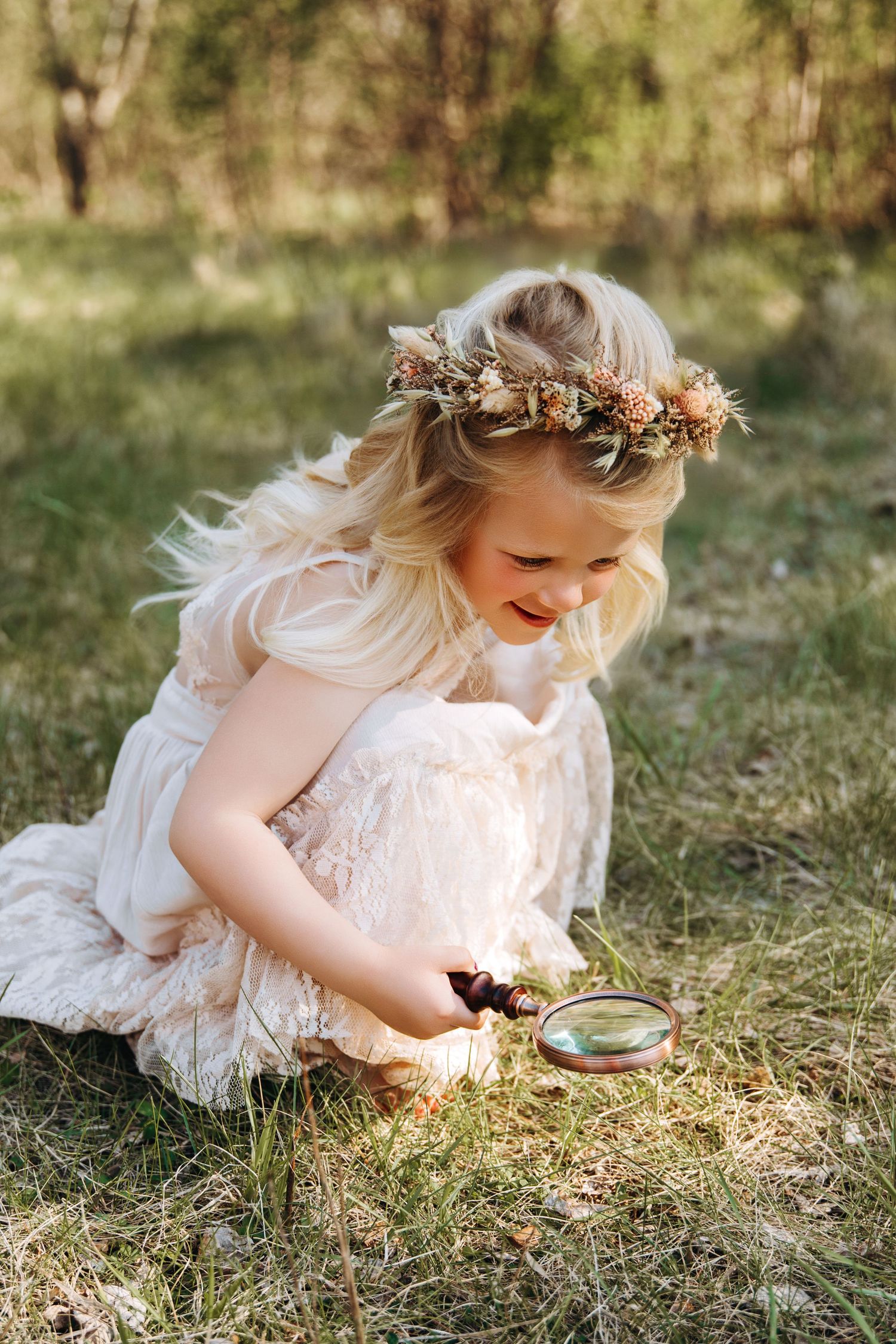 Child in white lace dress with flower crown explores nature using magnifying glass in sun-dappled outdoor setting.