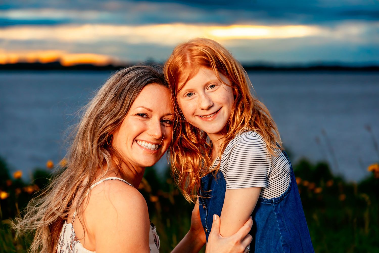 Two people smiling together at sunset by a lake with golden light illuminating the scene.