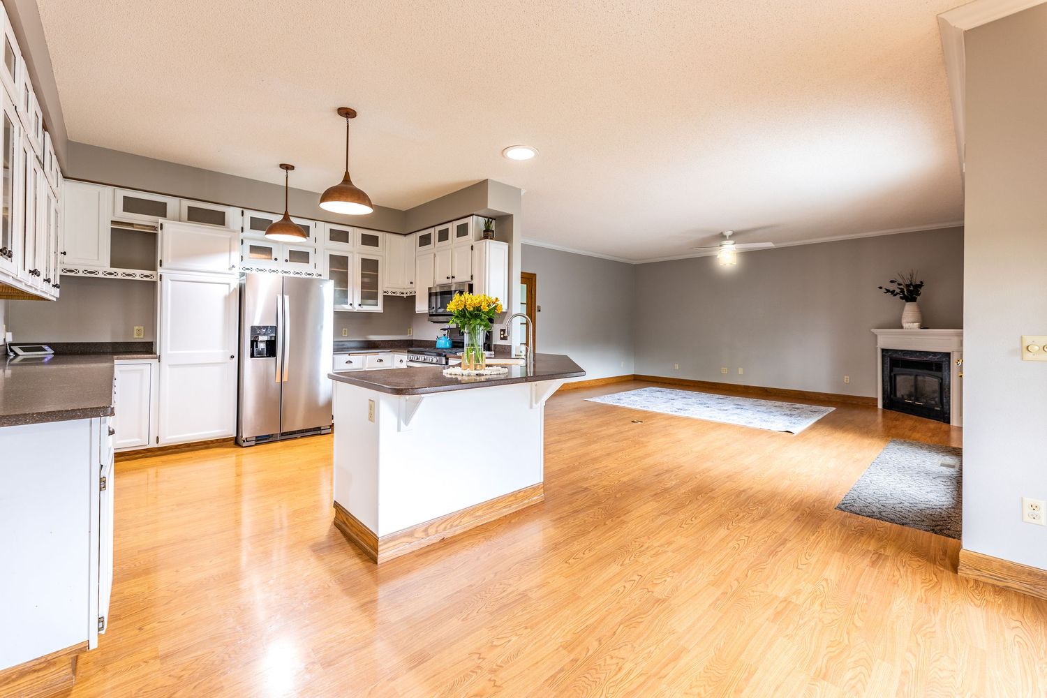 Open-concept kitchen with white cabinets, stainless steel fridge, island, pendant lights, and hardwood floors leading to living area.