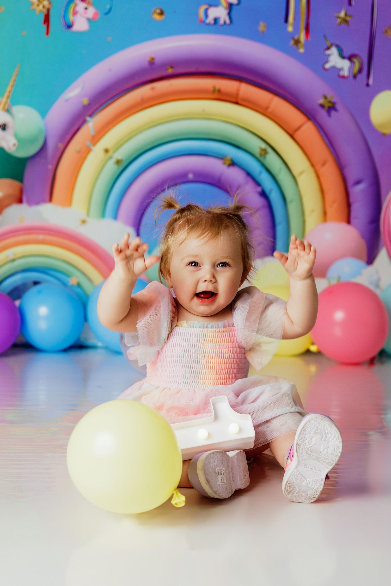 Excited baby girl in pink dress laughs with hands raised among colorful balloons and unicorn rainbow backdrop.