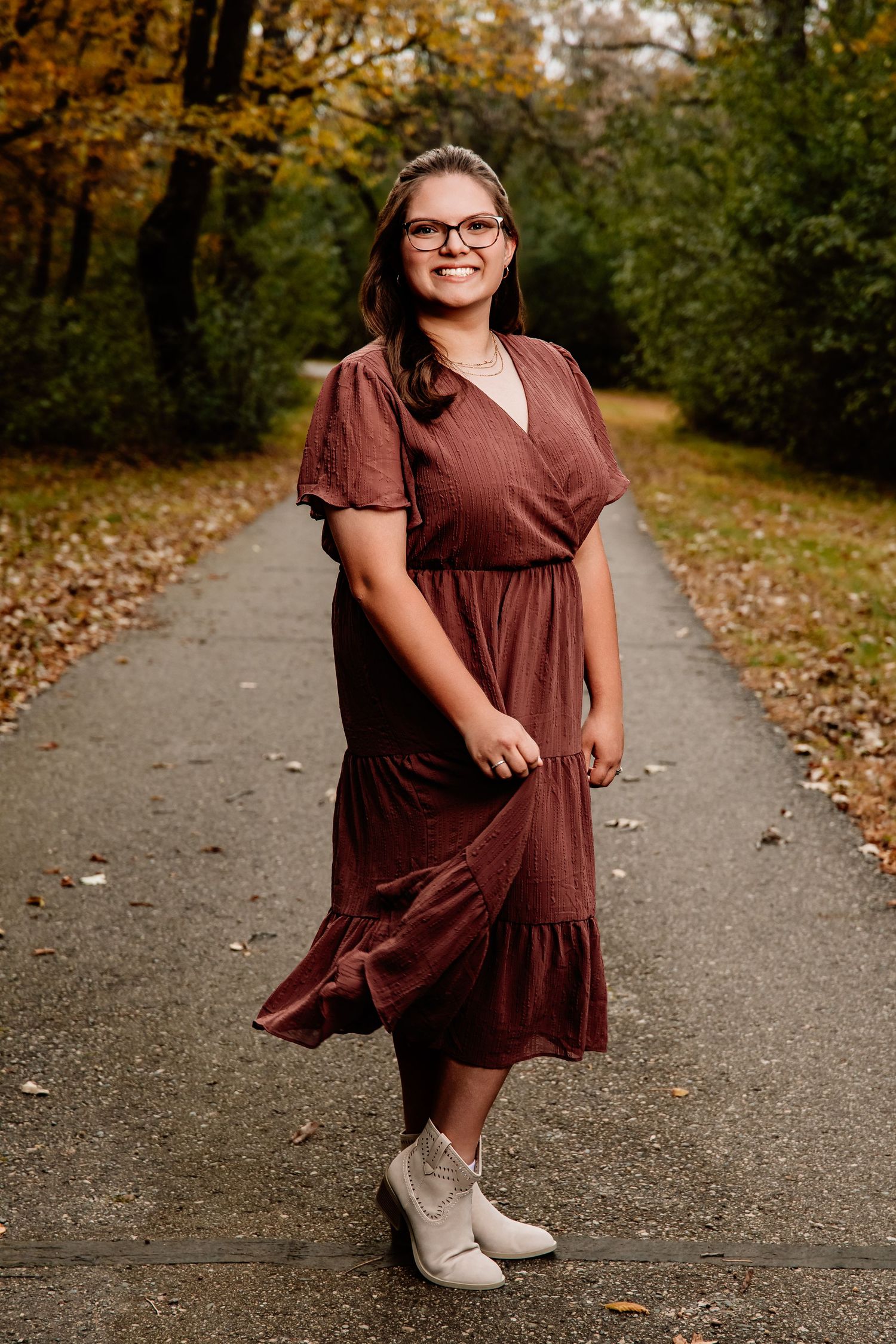 A person in a brown dress twirls playfully on a leaf-covered path in an autumn forest setting.
