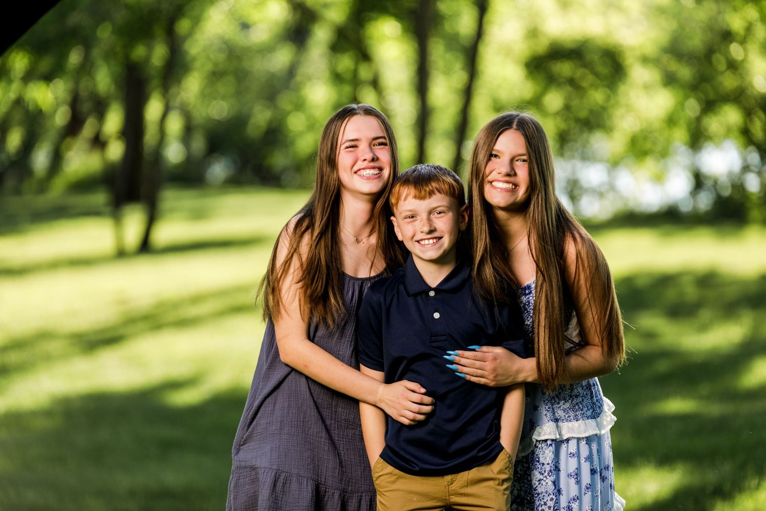 Three siblings smiling together outdoors, two girls with long hair hugging a young red-haired boy.