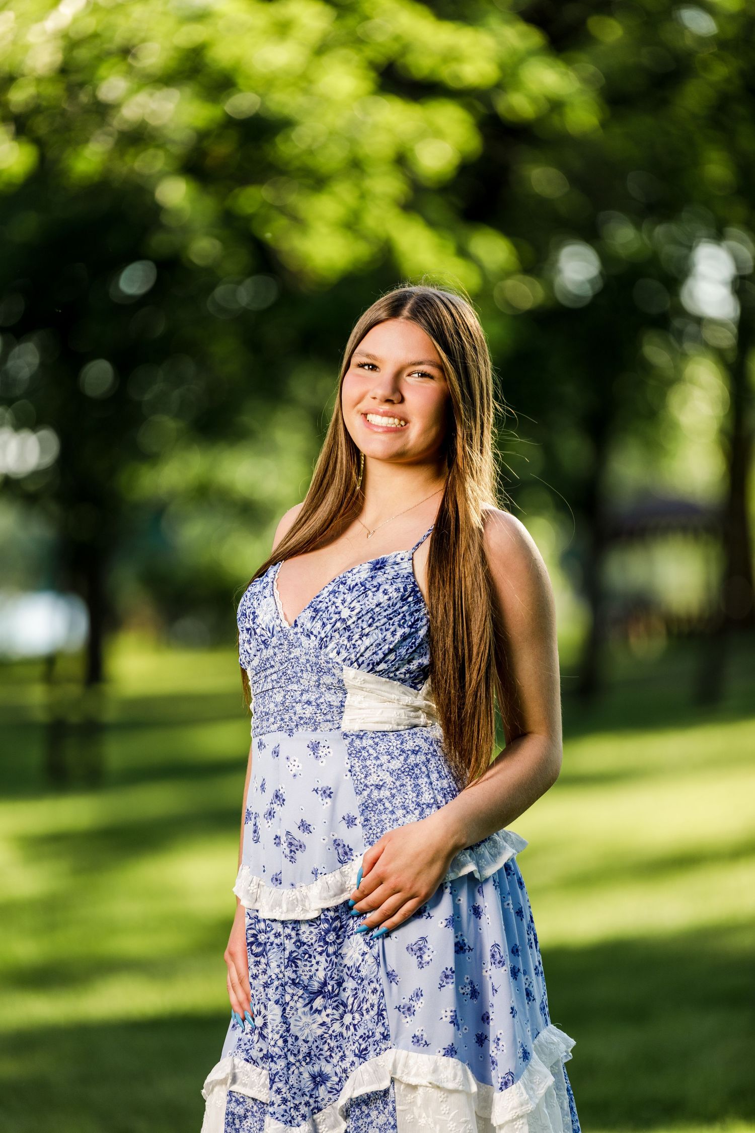 Smiling teenage girl in a blue floral sundress standing in a sunny green park.