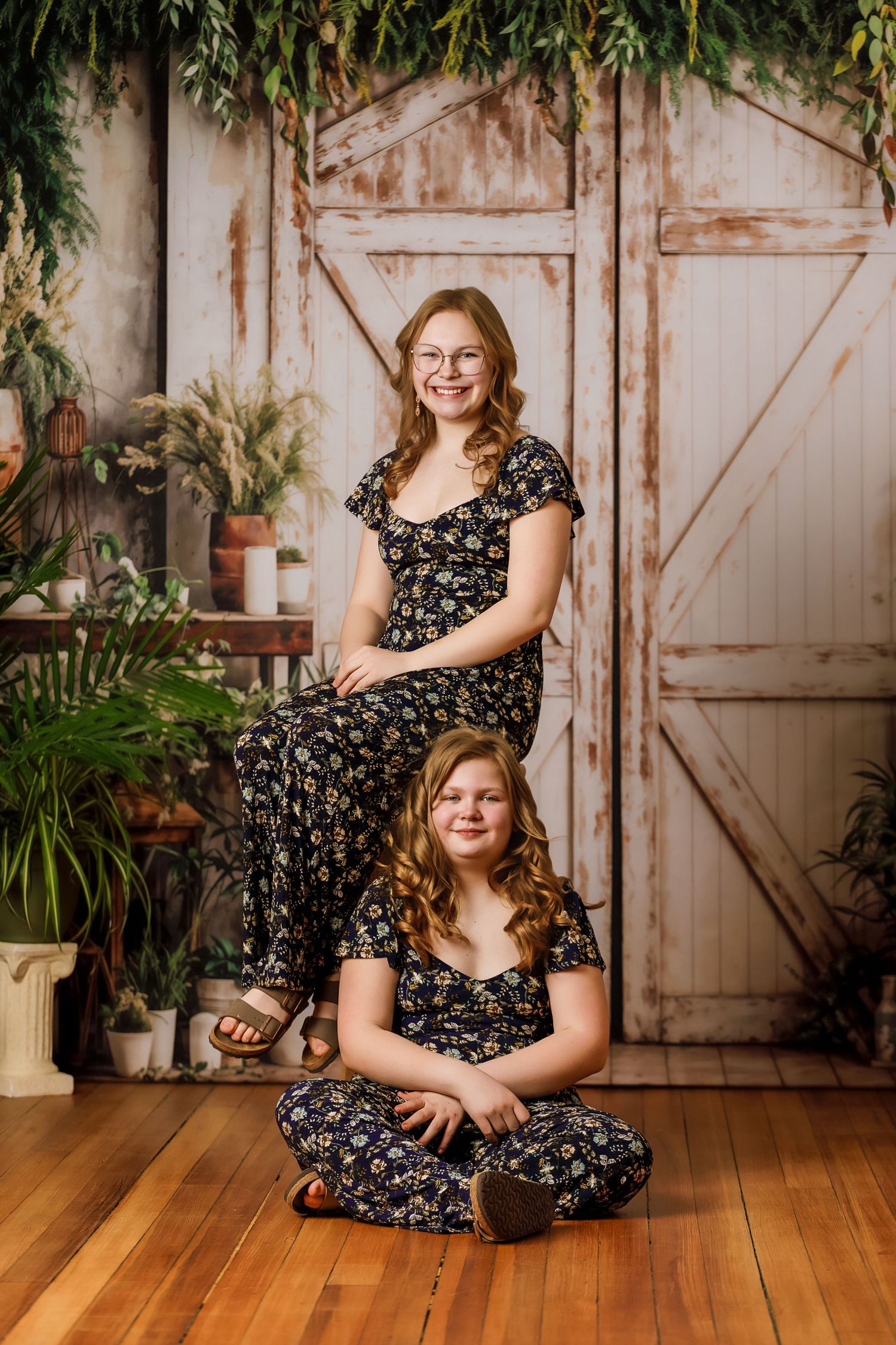 Two girls in matching floral jumpsuits pose in front of a rustic barn door backdrop with lush greenery.