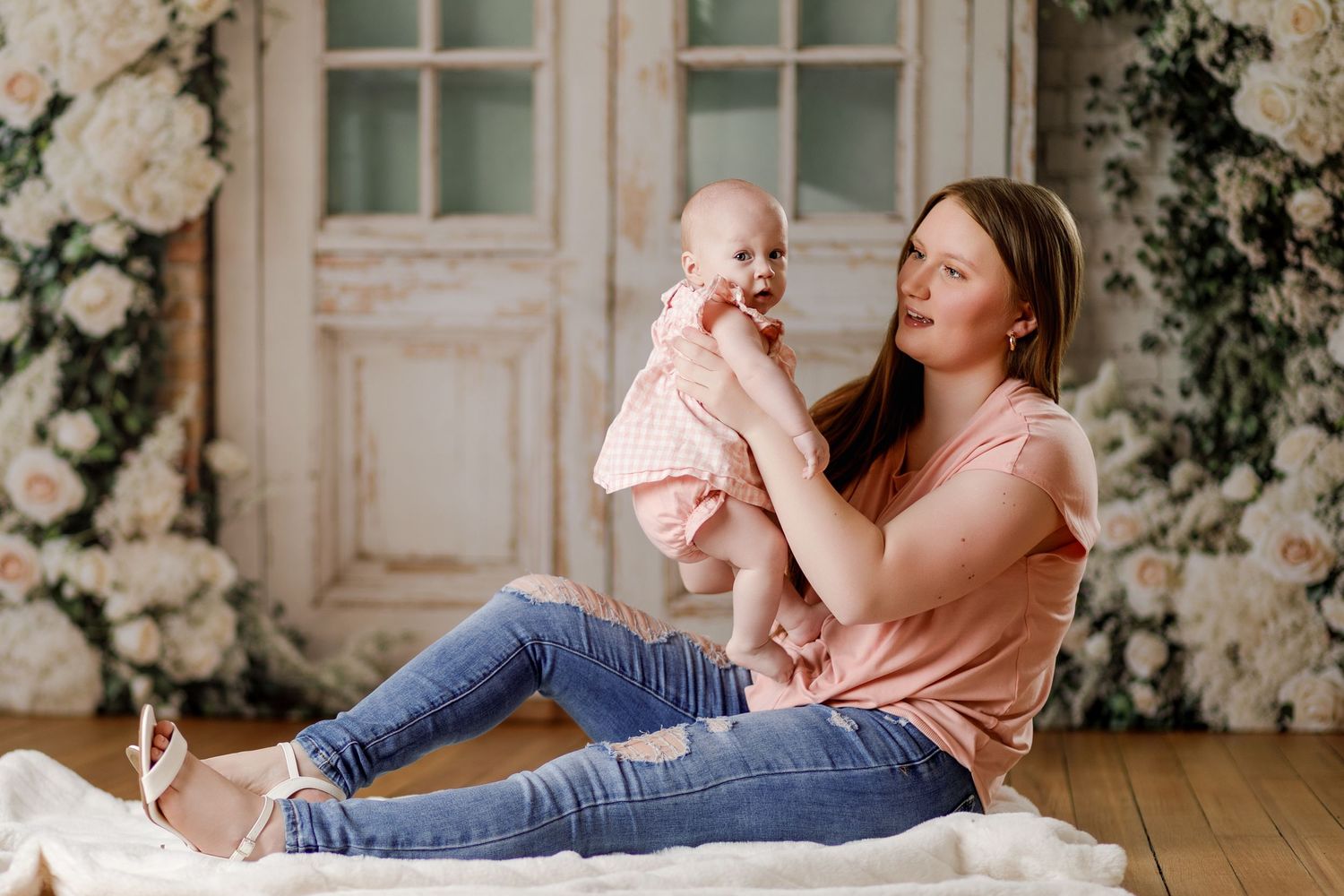 Young mother in pink shirt sits on floor holding her smiling baby in a floral studio backdrop.
