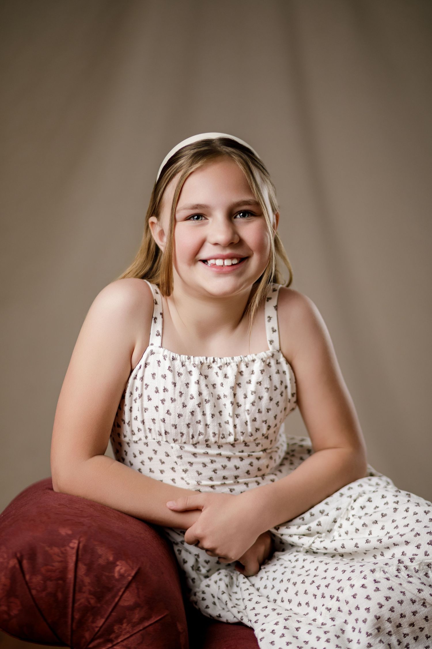 Smiling young girl in white floral dress and headband sitting gracefully in a studio portrait setting.