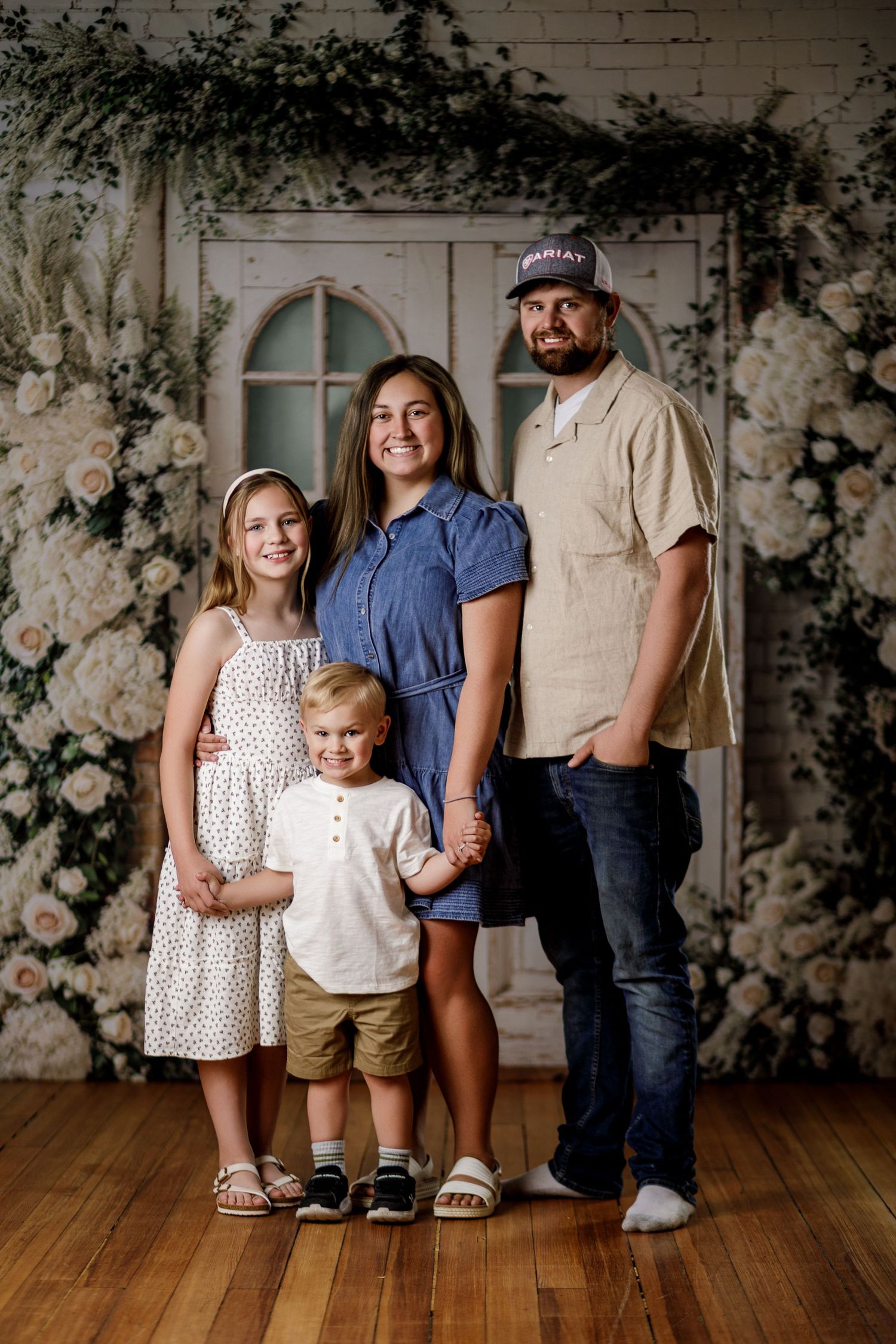 Family of four posing for a portrait with floral backdrop, featuring two parents and two children smiling together.