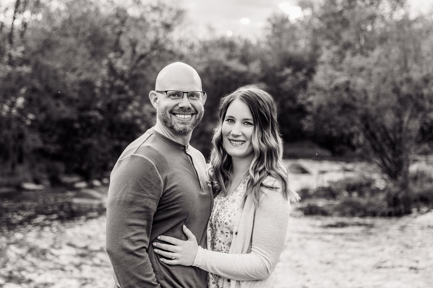 Black and white photo of a smiling couple embracing outdoors with trees in the background.