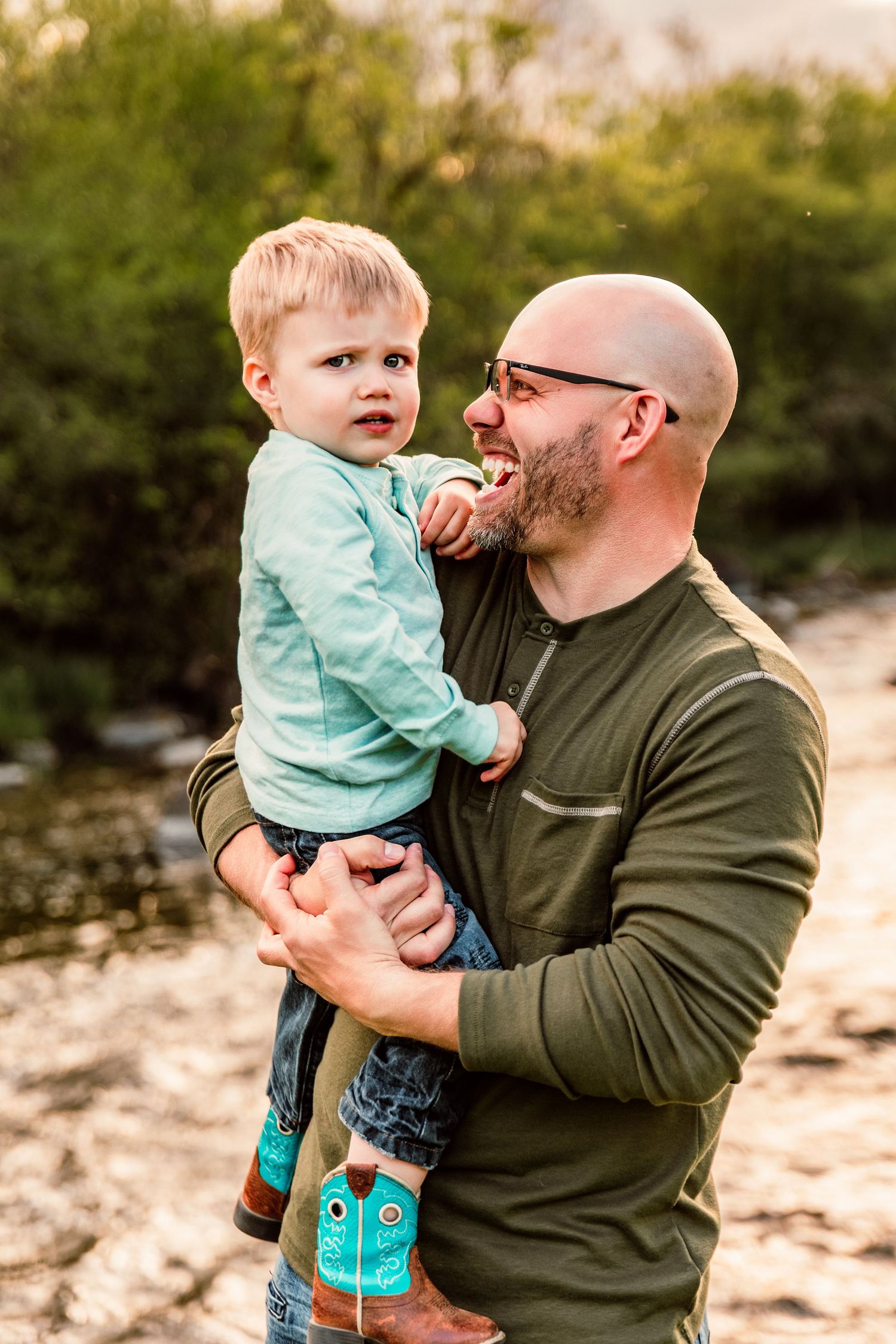 Bald father with beard holds young blonde toddler boy outdoors near creek, both wearing green shirts.