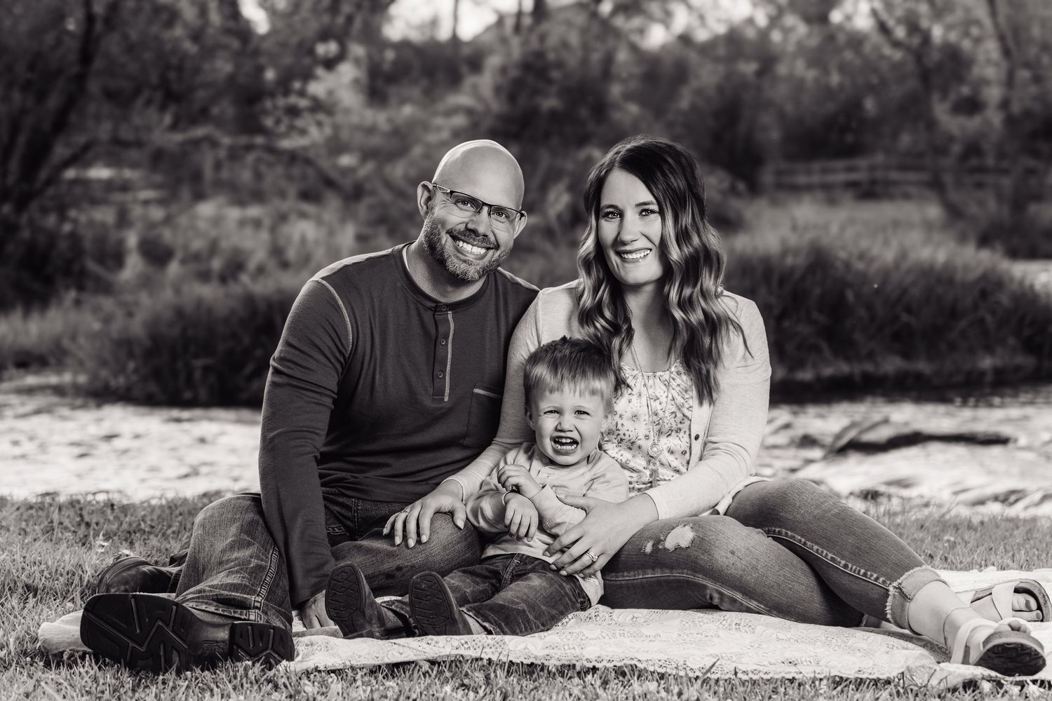 Black and white family portrait of smiling couple with baby sitting on blanket outdoors in natural setting.