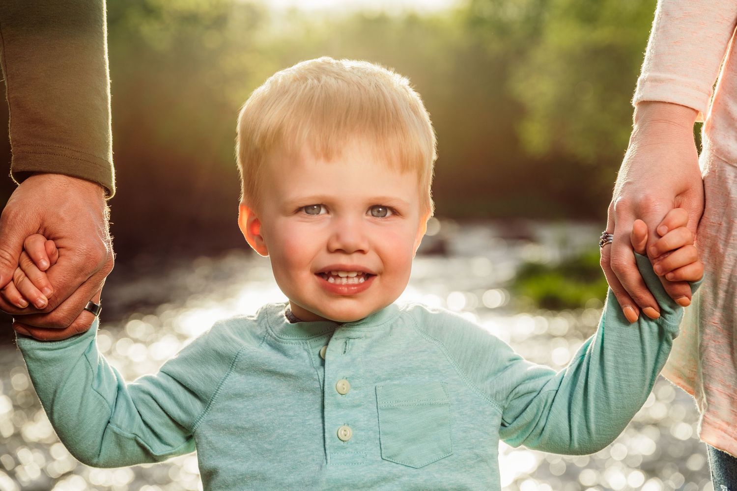 Happy toddler boy with blonde hair holding parents hands outdoors in golden sunlight near a river.