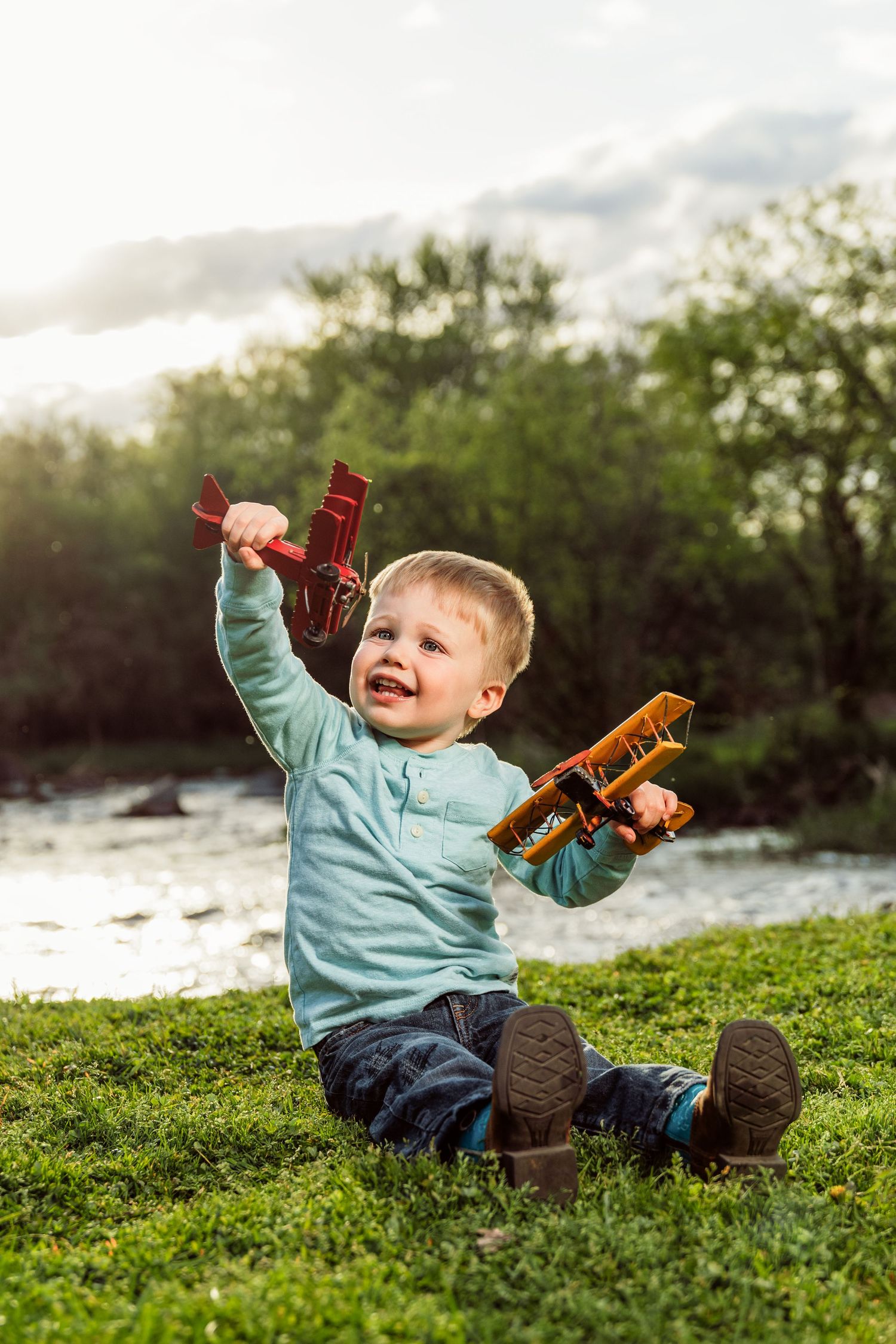 Happy toddler boy sitting on grass playing with two wooden toy airplanes outdoors near a river on a sunny day.