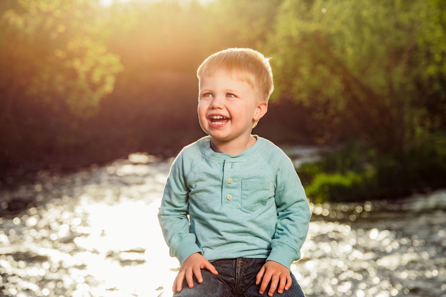 Happy toddler boy laughing outdoors by a sunlit river, wearing a teal shirt and jeans on a sunny day.