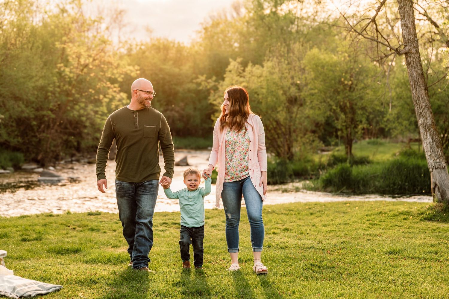 Family of three walking hand-in-hand on green grass near a river during golden hour sunset outdoors.