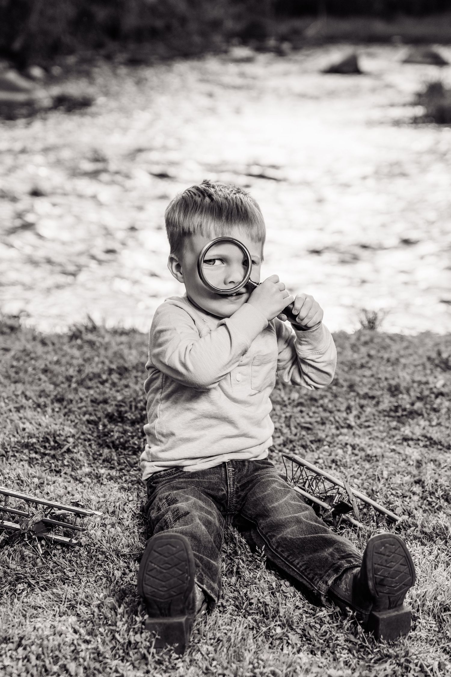 Black and white photo of a young boy sitting on grass, holding a magnifying glass up to his eye outdoors.